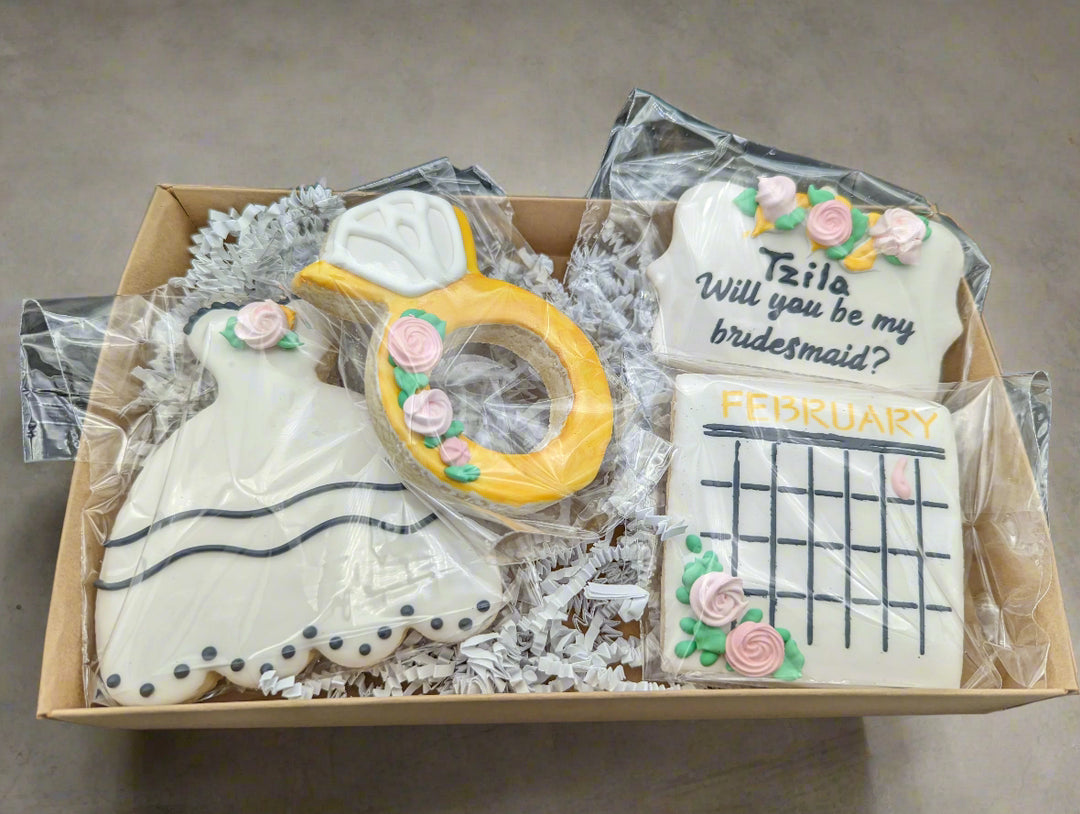 Box of cookies shaped like a wedding dress, ring, and calendar with 'Will you be my bridesmaid?' question.